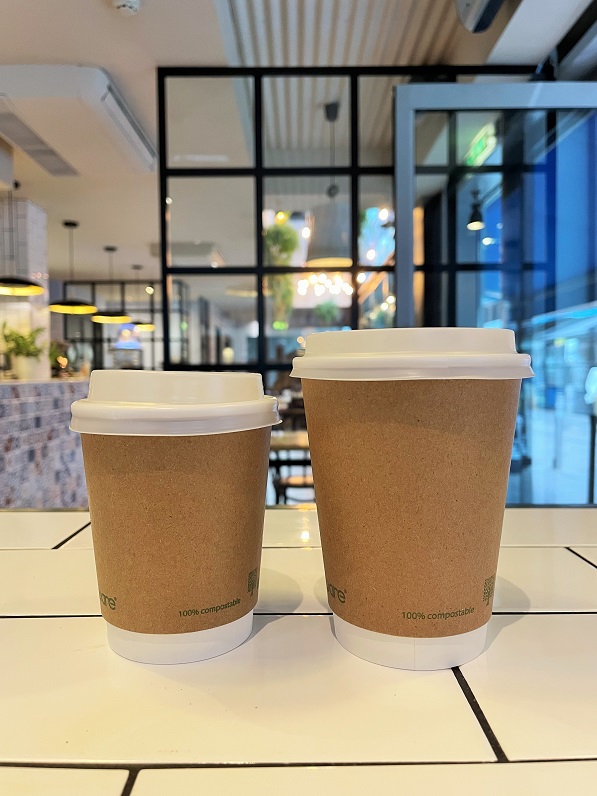 Foreground is large and small brown, recycable coffee cups with white lids on a white tiled counter at Jay Kays coffee shop in Dublin city centre. Background shows the full length of Jay Kays