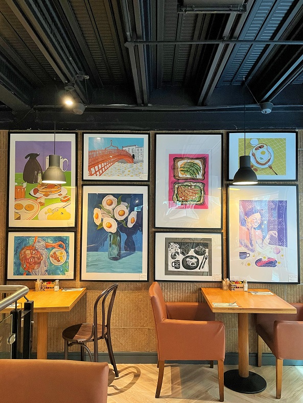 Modern art wall in the comfortable upstairs lounge of Jay Kays Café, showing eight paintings of paintings with an egg and breakfast theme. In front of the wall and below the paintings are tables and chairs