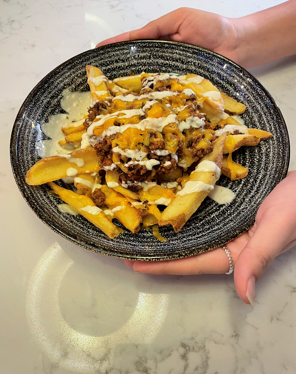 A blue and white speckled striped plate being held by two hands on a white marble counter. The plate shows a portion of Jay Kay's Topped with house beef chilli, melted cheese and jalapeño and lime mayo drizzled back and forth across the dish