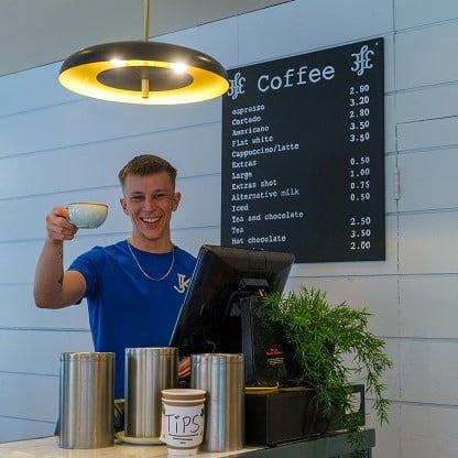 Young waiter standing behind the coffee counter in Jay Kay's Cafe. In front of him is a cash desk with a green fern on it. There are three metal cylindrical containers and a coffee cup with a handmade tips sign. In the background is a black sign with Coffee as the white heading and the full coffee menu below